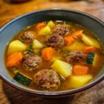 Close-up of a delicious bowl of albondigas soup featuring savory meatballs, chunks of potato, carrots, and zucchini in a rich broth.