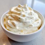 A close-up of thick, creamy homemade garlic aioli, swirled high in a small white bowl, showing flecks of pepper and minced garlic.