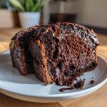 Two thick slices of rich, dark hot fudge brownie bread drizzled with melted chocolate sauce on a white plate.