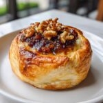 Close-up of a small, individual baked brie wrapped in flaky pastry, topped with a sticky glaze and walnuts.
