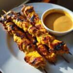 Four perfectly grilled, golden-yellow chicken satay skewers resting on a white plate next to a small bowl of peanut sauce.