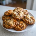 A generous pile of freshly baked kitchen sink cookies loaded with chocolate chips and pretzel pieces on a white plate.