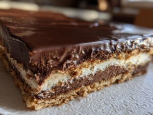 A close-up view showing the layers of a delicious eclair cake, featuring a graham cracker crust, cream filling, chocolate pudding, and shiny chocolate glaze.