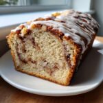 A close-up of a moist apple bread loaf, sliced to show cinnamon swirl and walnuts, topped with white icing.