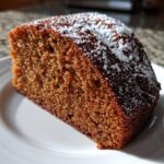 Close-up of a moist, dark slice of applesauce cake dusted with powdered sugar on a white plate.