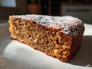 Close-up of a moist, brown slice of applesauce cake dusted lightly with white powdered sugar on a white plate.
