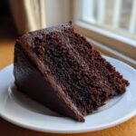 Close-up of a thick slice of moist chocolate cake covered in dark, glossy chocolate frosting on a white plate.