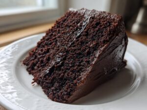 A close-up of a rich, dark slice of moist chocolate cake covered in thick chocolate frosting on a white plate.