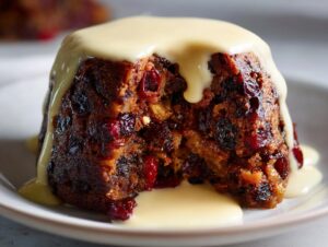 Close-up of a rich, dark plum pudding with visible dried fruit, topped with thick vanilla custard sauce.