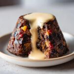 Close-up of a rich, dark plum pudding topped with thick, creamy custard sauce dripping down the side.