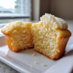 Close-up of a vanilla cupcake cut in half showing its soft, moist crumb and a swirl of white frosting.