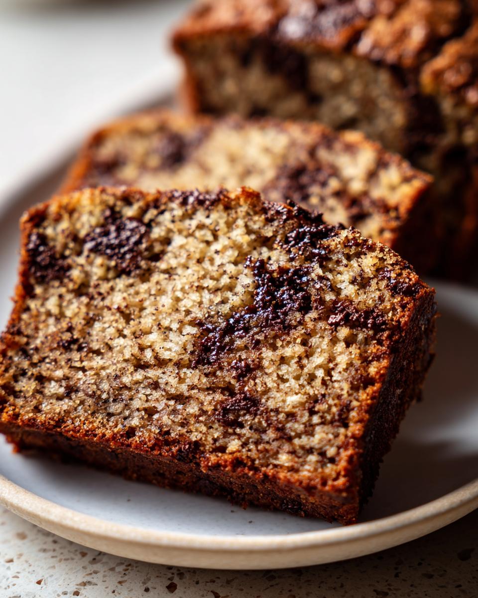 Close-up of a moist slice of vegan banana bread featuring dark chocolate swirls on a light plate.