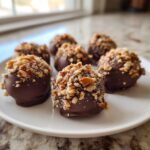 Close-up of several chocolate-dipped pecan pie balls generously topped with chopped nuts on a white plate.
