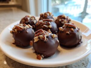 Close-up of several chocolate-covered pecan pie balls topped with chopped pecans, served on a white plate.