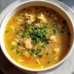 Close-up of a steaming bowl of golden penicillin soup topped with grated cheese, black pepper, and fresh parsley.