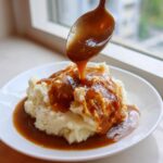 A spoonful of rich, brown chicken gravy is being poured over a mound of creamy mashed potatoes on a white plate.