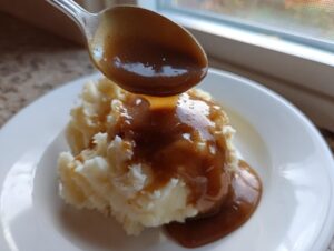 A spoonful of rich brown chicken gravy being poured over a mound of fluffy mashed potatoes on a white plate.