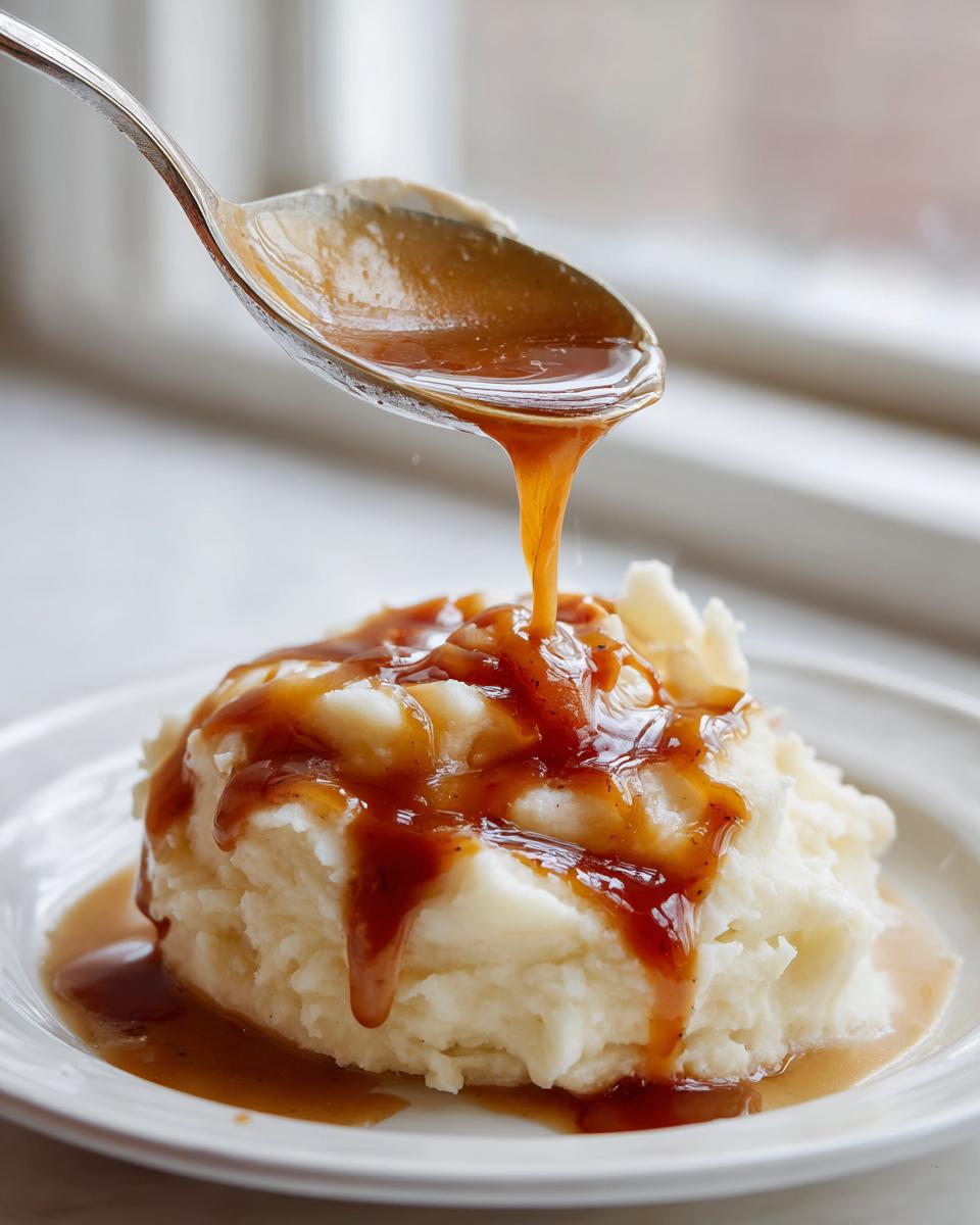 A spoonful of rich, brown chicken gravy is being poured over a mound of fluffy mashed potatoes on a white plate.