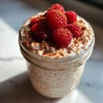 Close-up of creamy overnight oats topped with fresh raspberries and cinnamon powder in a glass jar.