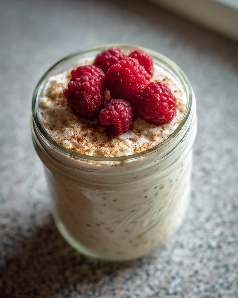 Close-up of creamy overnight oats topped with fresh raspberries and cinnamon in a glass jar.
