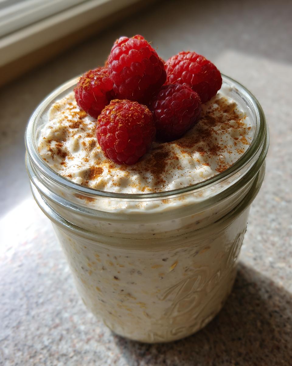 Close-up of creamy overnight oats in a glass jar, topped with fresh raspberries and cinnamon.