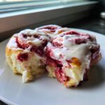 Two delicious red velvet cinnamon rolls covered in thick white cream cheese icing on a white plate.