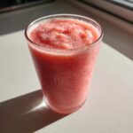 A close-up of a frosty, bright pink watermelon smoothie served in a clear glass, glistening in sunlight.