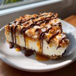A decadent slice of Samoa Cheesecake topped with toasted coconut, caramel, and chocolate drizzle.