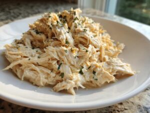 A close-up of shredded slow cooker garlic parmesan chicken, creamy and topped with green herbs, served on a white plate.