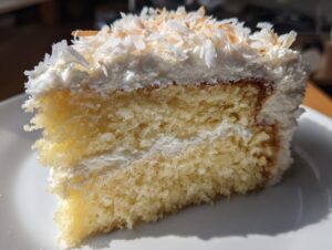 Close-up of a moist slice of coconut cake layered with white frosting and topped with toasted coconut flakes.