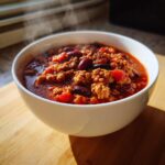 A close-up of a steaming white bowl filled with rich, hearty high protein chili featuring ground meat and kidney beans.