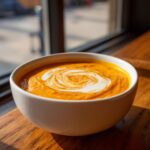 A close-up of vibrant orange butternut squash soup in a white bowl, topped with a decorative swirl of cream.