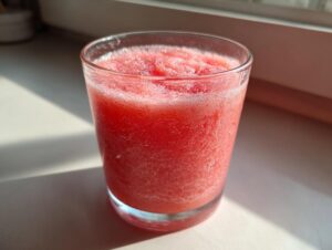 Close-up of a bright pink, icy watermelon smoothie served in a clear glass, catching sunlight.