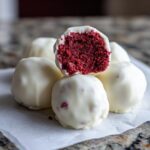 Close-up of several white chocolate covered red velvet cake balls, one cut in half revealing the bright red interior.