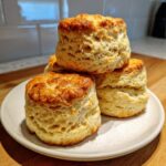 A stack of four tall, golden brown, freshly baked scones with flaky sides, resting on a light-colored plate.