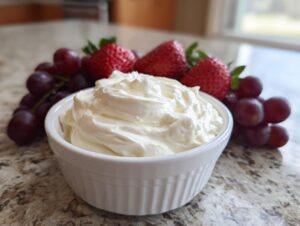 A white bowl filled with creamy fruit dip, surrounded by fresh red grapes and strawberries.