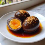 Three deeply colored marinated eggs on a white plate, one cut in half showing a jammy yolk, topped with sesame seeds.