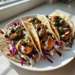 Close-up of three perfectly grilled shrimp taco shells filled with cabbage slaw, topped with seasoned shrimp, crema, and herbs.