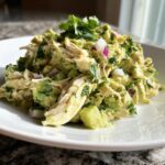 A mound of creamy avocado chicken salad mixed with chunks of avocado, red onion, and cilantro, served on a white plate.