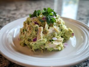 A scoop of creamy avocado chicken salad mixed with shredded chicken and red onion, topped with fresh parsley.