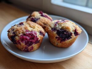 Three freshly baked berry protein muffins with a sugary crust served on a white plate.