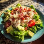 A vibrant serving of blt chicken salad piled high with shredded chicken, bacon bits, avocado chunks, and tomatoes on lettuce.