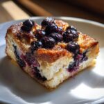 A golden brown slice of blueberry french toast casserole topped with glazed blueberries, served on a white plate.