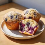 Three blueberry protein muffins on a white plate, one cut in half showing the moist interior packed with blueberries.