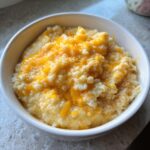 A close-up of a white bowl filled with creamy cheese grits topped with melted, bright orange cheddar cheese.