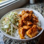 A white bowl filled with white rice, topped with saucy buffalo chicken pieces and a side of creamy coleslaw, making a perfect buffalo chicken bowl.