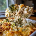 A wooden spoon lifts a cheesy scoop of chicken and broccoli rice casserole, showing long cheese pulls.