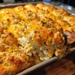A wooden spoon lifts a portion of cheesy chicken cobbler, showing fluffy biscuits and melted cheddar topping.