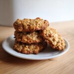 A stack of four chewy banana oatmeal cookies resting on a small white plate on a wooden surface.
