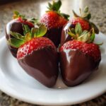 Five glossy chocolate covered strawberries with bright red tops and green stems arranged on a white plate.